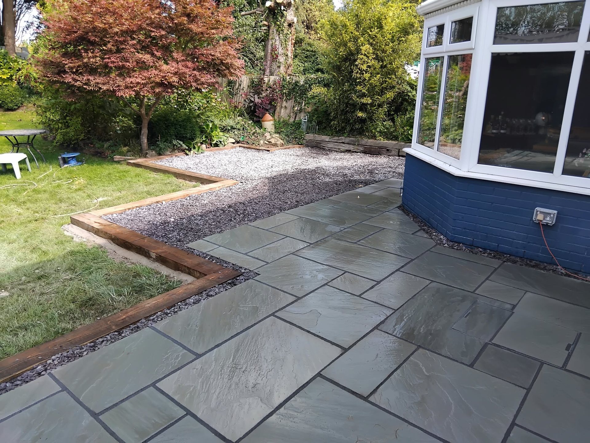Grey sandstone patio with timber sleeper edging and slate chippings beside a lawn